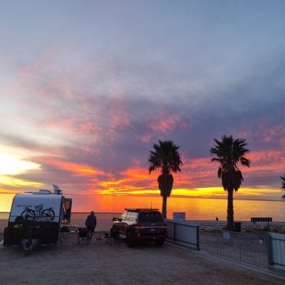 Caravan and vehicle parked at Smoky Bay beachfront during sunset with palm trees and vibrant sky.