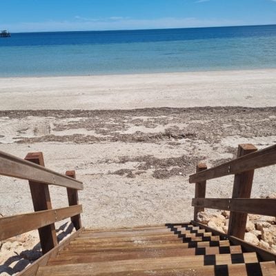 Wooden staircase leading down to a sandy beach with clear blue ocean and cloudless sky.