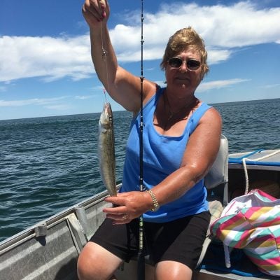 Woman fishing from a boat holding a caught fish with water and shoreline in the background